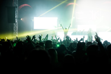 People with hands up in the air on the dance floor during a concert. A great glow covers the singer and the artistic animation on the stage. Laser and lights illuminate the party