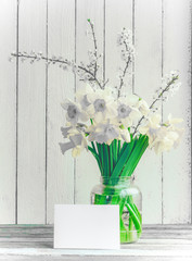Bouquet of daffodilsin and blossoming plum branch a glass jar with a mock up blank greeting card on a wooden table in the background of the white wooden wall with copy space