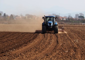 Obraz premium Tractor plows the dry agricultural field raising dust, in a sunny morning of early springtime