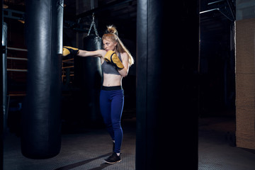 Young sportswoman boxer practicing near bag, ring in gym.