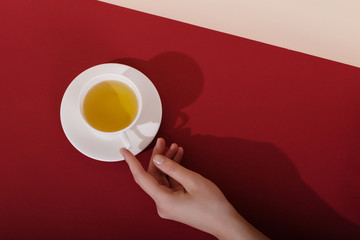 Ceramic cup with tea, on a red background. The harsh shade of the sun, the art concept of the morning and the charge of energy. Girl hand reaches for the mug.