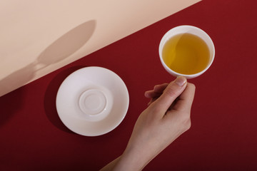Ceramic cup with tea, on a red background. The harsh shade of the sun, the art concept of the morning and the charge of energy. Girl hand reaches for the mug.