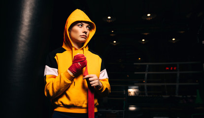 Sports girl boxer in yellow sweatshirt pulls red bandages on her hands.