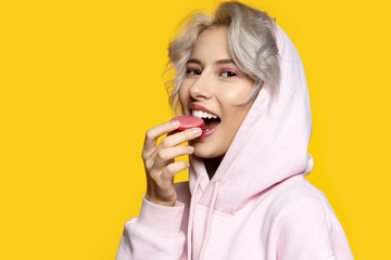 Portrait of sweet lady eating pink macaron and looking at camera. Pretty blonde girl and tasty cake. Food and pleasure concept. Isolated on yellow background