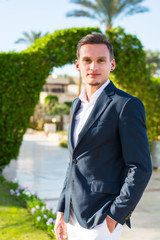 Young handsome man in a white shirt, jacket and white trousers at the palm trees background on a beach.