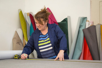 Older woman in a furniture factory is measuring and marking a grey material for the sofa with a chock