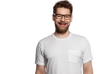 Portrait of happy young bearded man smiling while wearing blank shirt and posing on white background. Cheerful brunette guy with attractive appearance. Fashion and lifestyle concept