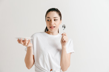 Young fun crazy brunette housewife with spoon isolated on white background. Housekeeper woman holding white empty plate. Copy space advertisement.