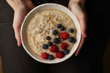 Plate with porridge in hand on dark background, top view. Healthy Breakfast with raspberries and blueberries, chia seeds, maple syrup.