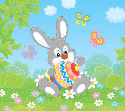 Little Easter Bunny sitting with a colorfully decorated big egg among flowers and flittering butterflies on a green lawn on a sunny spring day, vector illustration in a cartoon style
