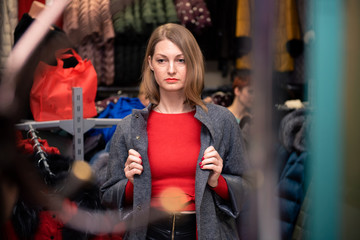 Fototapeta na wymiar Young adult girl in a clothing store in a red sweater trying on a gray coat against the background of hanging clothes