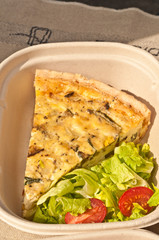 Top view, close up of a slice of french breakfast quiche, with lettece and cherry tomato, freshly baked in a cardboard container on display and for sale at a tropical farmers market