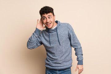 Young man over isolated wall listening to something by putting hand on the ear