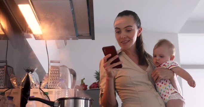 Woman holds child with one hand second phone cooks kitchen same time