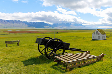 Fototapeta na wymiar Green meadow on Iceland with old sled, cart and barn with snow mountains on background