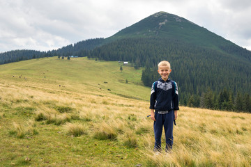 Fototapeta na wymiar Young happy smiling child boy with backpack standing in mountain grassy valley on background of summer landscape, woody mountain view. Active lifestyle, adventure and weekend activity concept.