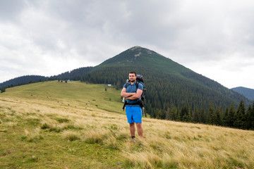 Young handsome man with backpack standing in mountain grassy valley on copy space background of summer woody mountain peak and blue sky. Active lifestyle, sports and recreation concept
