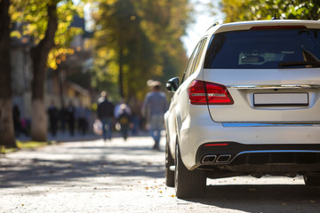 Back view of white car parked on city pedestrian zone on background of blurred silhouettes of people walking along green sunny summer alley. Modern city lifestyle concept.