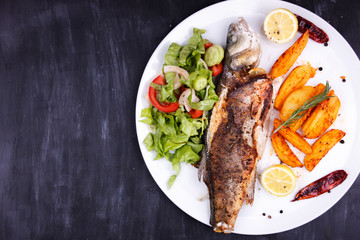Fried sea bass with lemon, potatoes, salad and spices on a black wooden table