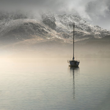Stunning landscape image of sailing yacht sitting still in calm lake water with mountain looming in background during Autumn Fall sunrise