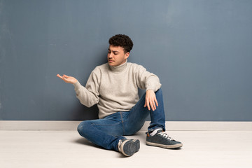 Young man sitting on the floor holding copyspace with doubts