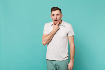 Portrait of shocked bewildered young man in casual clothes put hand prop up on chin isolated on blue turquoise wall background in studio. People sincere emotions lifestyle concept. Mock up copy space.