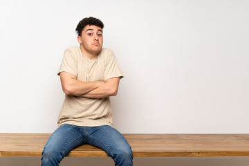 Young man sitting on table making doubts gesture while lifting the shoulders