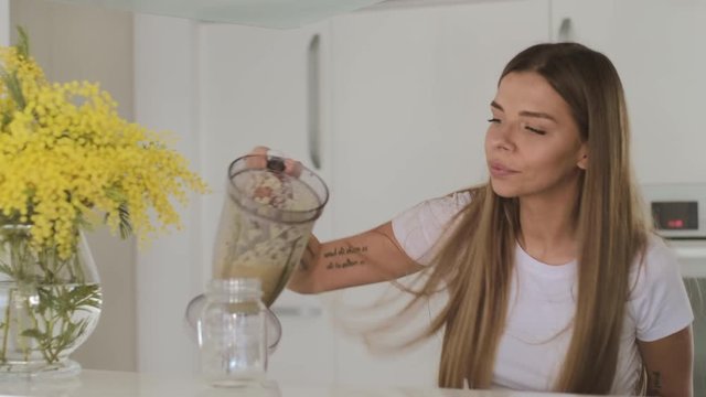 Young blond woman in white t-shirt prepares fruit smoothie in blender at home in the kitchen
