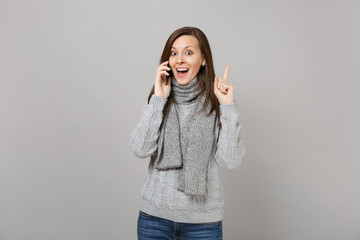 Excited woman in gray sweater, scarf holding index finger up with great new idea, talking on mobile phone isolated on grey background. Healthy fashion lifestyle, people emotions, cold season concept.