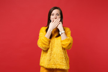 Portrait of shocked scared young woman in yellow fur sweater covering mouth with hands isolated on bright red wall background in studio. People sincere emotions, lifestyle concept. Mock up copy space.