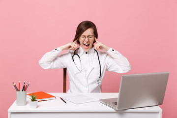 Tired mad female doctor sits at desk work on computer with medical document in hospital isolated on pastel pink wall background. Woman in medical gown glasses stethoscope. Healthcare medicine concept.