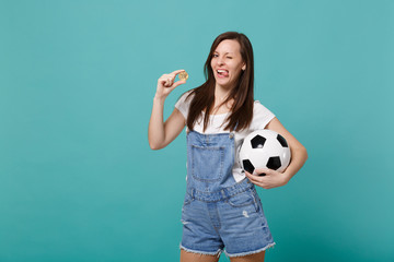 Cheerful blinking girl football fan support favorite team with soccer ball, bitcoin future currency showing tongue isolated on blue turquoise background. People emotions, sport family leisure concept.