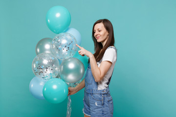 Fototapeta premium Pretty young woman in denim clothes celebrating, holding and pointing index finger on colorful air balloons isolated on blue turquoise wall background. Birthday holiday party, people emotions concept.