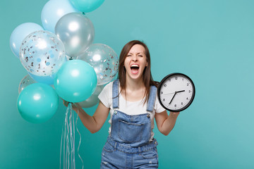 Crazy screaming young woman in denim clothes holding round clock while celebrating with colorful...