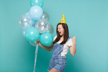 Overjoyed young woman in birthday hat screaming doing winner gesture, celebrating, holding colorful air balloons isolated on blue turquoise background. Birthday holiday party, people emotions concept.