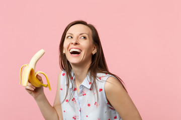Laughing young woman in summer clothes looking up holding fresh ripe banana fruit isolated on pink pastel wall background in studio. People vivid lifestyle relax vacation concept. Mock up copy space.
