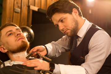 Attractive young professional barber shaving his client with a razor working at the barbershop copyspace profession occupation job barbering service concentration hipster lifestyle fashion styling.