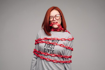 Studio portrait of a girl entangled in a chain. A red-haired woman with brown hair tries to get rid of the chain. The concept of a life-like situation. Isolated on a gray background.