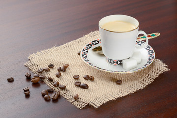 Coffee in porcelain cup with saucer and golden spoon on a jute napkin. Vintage coffee set on wooden table with sugar and coffee beans