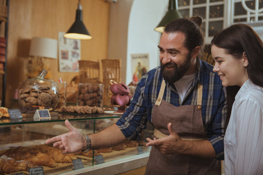 Bearded mature baker helping his customer choosing pastry from the display. Beautiful happy woman shopping at the bakery. Cheerful baker working at his store, selling delicious food, copy space