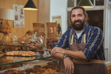 Handsome mature bearded male baker smiling to the camera proudly, working at his bakery shop, copy space. Friendly baker with manbun hairdo leaning on the showcase at his cafe