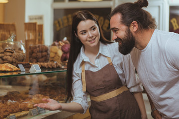 Attractive female baker wearing apron, offering her customer pastry on the display, copy space. Handsome mature long haired man smiling, buying delicious desserts at the bakery