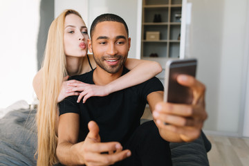 Lovely couple moments of handsome guy and young pretty woman with long blonde hair having fun, making selfie on phone. Chilling on bed at home in modern apartment, kissing, in love