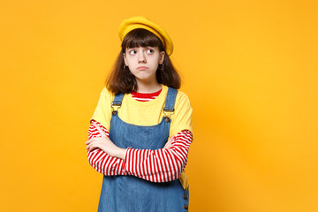 Portrait of pensive girl teenager in french beret, denim sundress holding hands crossed, looking up isolated on yellow wall background. People sincere emotions, lifestyle concept. Mock up copy space.