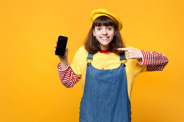 Pretty girl teenager in french beret pointing index finger on mobile phone with blank empty screen isolated on yellow wall background. People sincere emotions, lifestyle concept. Mock up copy space.