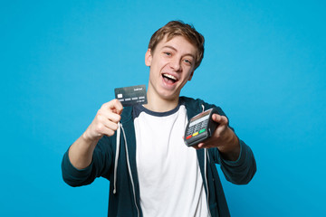 Funny young man holding wireless modern bank payment terminal to process and acquire credit card payments isolated on blue background. People sincere emotions, lifestyle concept. Mock up copy space.