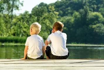 Two little boys are sitting on the pier on the river bank. Concept of friendship and fraternity. Back view