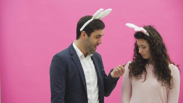 Young couple are beautiful on pink background. During this time, they are dressed in rabble ears. Looking at each other. A man shows carrots, his wife does not like the size of carrots and she throws