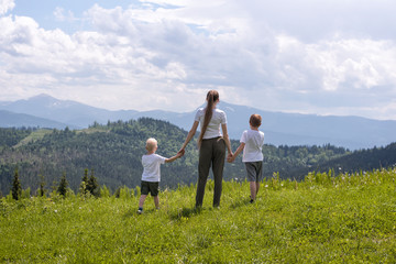 Naklejka premium Mother and two little sons stand holding hands on a green field against a background of forest, mountains and sky with clouds.