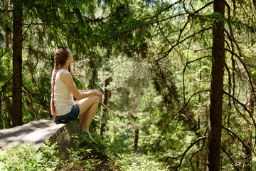 Dreaming beautiful girl sitting on a stone surrounded by coniferous forest on a sunny day